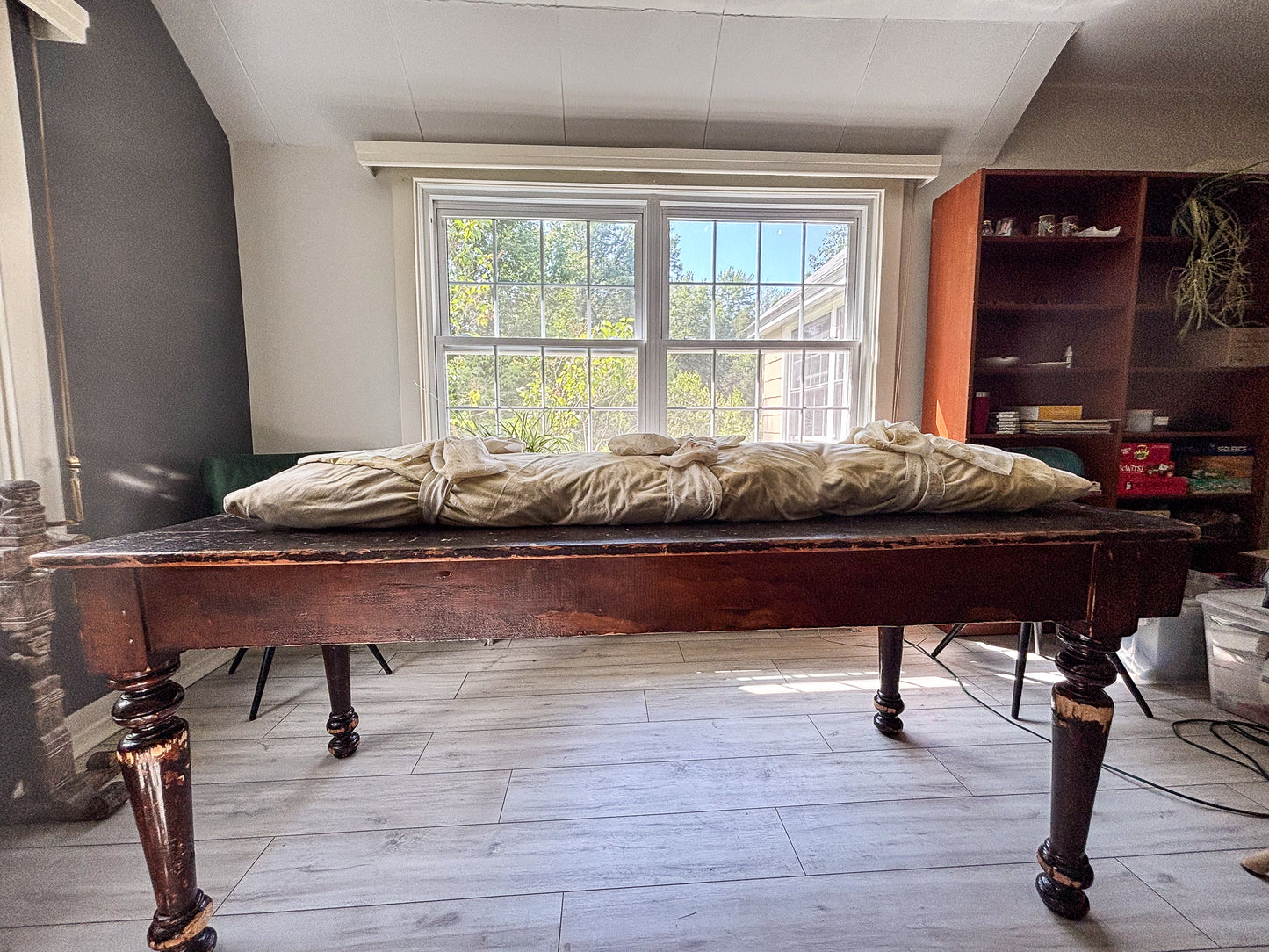 Wooden table with a floral-dyed burial shroud on top in a room with a window and shelves.