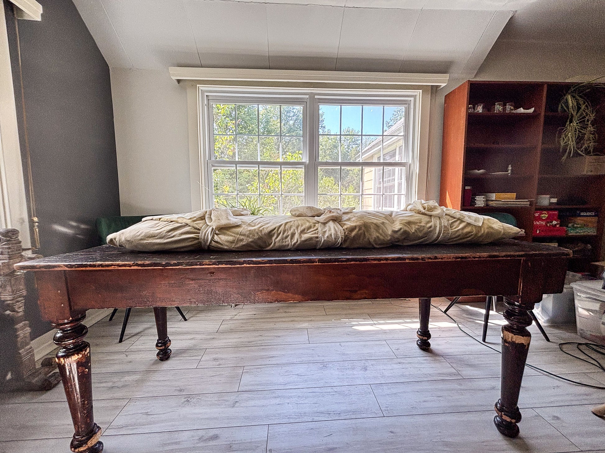 Wooden table with a floral-dyed burial shroud on top in a room with a window and shelves.
